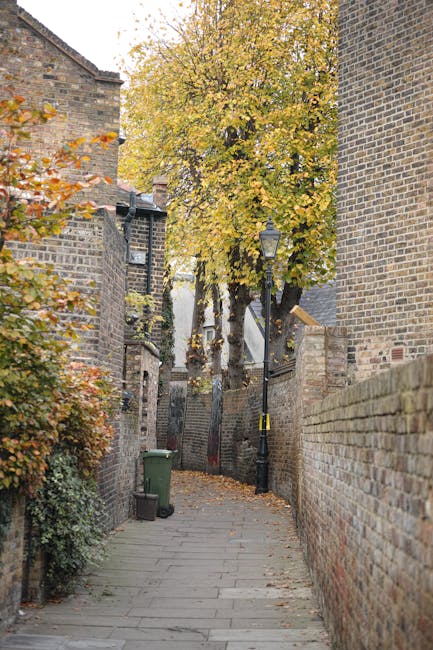 A narrow cobblestone alleyway enclosed by tall brick walls on both sides, with a large deciduous tree displaying yellow and orange autumn leaves positioned at the end of the passage. Two traditional b