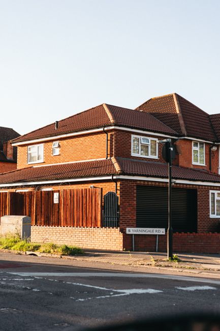 A two-storey brick house with a pitched red tile roof situated on a street corner, featuring white-framed windows on both levels and a black garage door on the ground floor. A wooden fence runs along