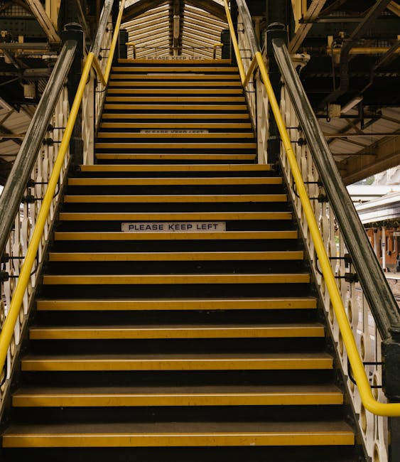 A narrow outdoor staircase with dark grey steps ascending between two tall, beige-painted building walls. The walls feature visible piping and electrical conduits, with some pipes mounted on the left 